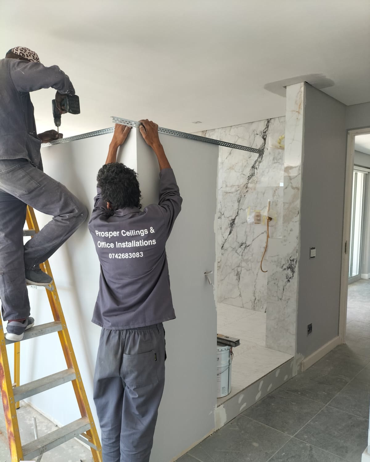 Installers in Prosper Ceilings workwear fixing a partition wall track beside marble-clad shower cladding